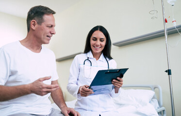 Fototapeta premium Happy cute confident woman doctor with a tablet in hands talks with the patient in the hospital ward