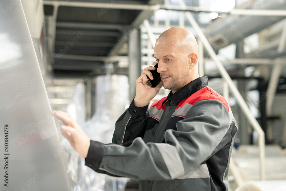 Poster bald mature worker with smartphone by ear pressing button on control panel