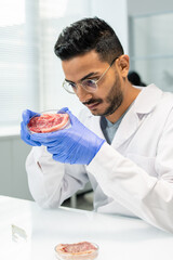 Young gloved researcher looking attentively at raw vegetable meat in petri dish