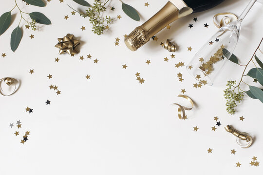 Happy New Year still life. Empty champagne glass, wine bottle with golden confetti stars and eucalyptus branches isolated on white table background. Celebration, party concept. Flat lay, top view.
