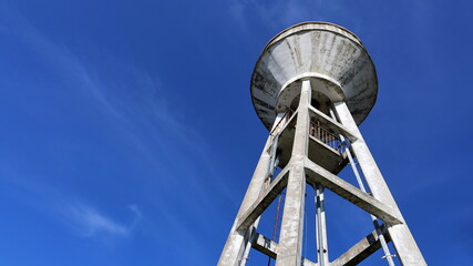 Concrete water tank on a tall tower. Large white storage tank for water supply systems in communities or workplaces. On a bright blue sky background with copy space. In view below. Selective focus