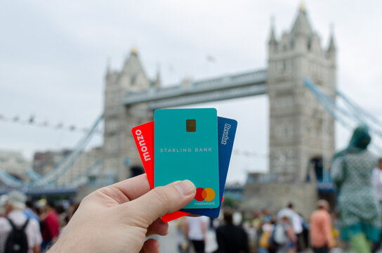 London / United Kingdom - July 6 2019: Revolut, Starling, Monzo Bank Cards Hold In Hand And London Tower Bridge At The Blurred Background. Conceptual: The Competing Fintech Companies Are London Based.