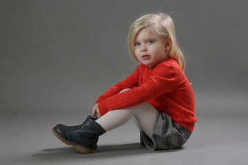 Fashion studio portrait of adorable 3 years old girl in red buttined sweater and plaid shorts on grey background.