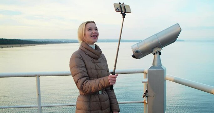 Cheerful Pretty Woman Talking on Video Communication via Smartphone While Standing on the Pier. Female Blogger, a Social Media Influencer, Talks to her Followers on Camera about her Journey.