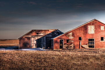 An old red barn at sunset in a rural setting. The barn in falling apart. There is a lot of red brick and boarded up windows. There is brown grass in the foreground.  - obrazy, fototapety, plakaty