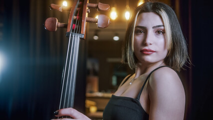 Beautiful young attractive woman in black dress posing for camera on stage with cello. Close-up