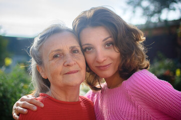 Senior mother with gray hair with her adult daughter looking at the camera in the garden and hugging each other during sunny day outdoors