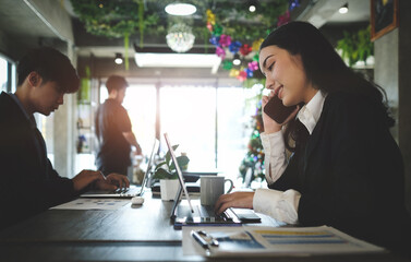 Young businesswoman in black suit talking on mobile phone and working with computer tablet while sitting  with her colleague in modern workplace.