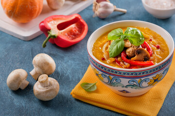 Pumpkin cream soup with basil, roasted mushrooms and red bell pepper on blue background