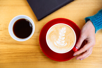 A mug of aromatic coffee cappuccino in a woman's hand on a table near a book. Next is another cup of coffee. Flat lay