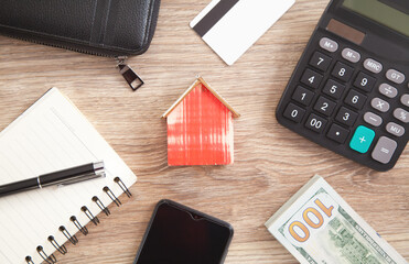 Wooden house model with a wallet, credit card, calculator, money, smartphone, pen and notepad.