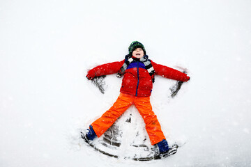 Cute little kid boy in colorful winter clothes making snow angel, laying down on snow. Active outdoors leisure with children in winter. Happy healthy child having fun and laughing outdoors