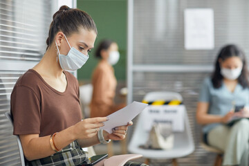 Side view portrait of young woman wearing mask and holding document while waiting in line in...