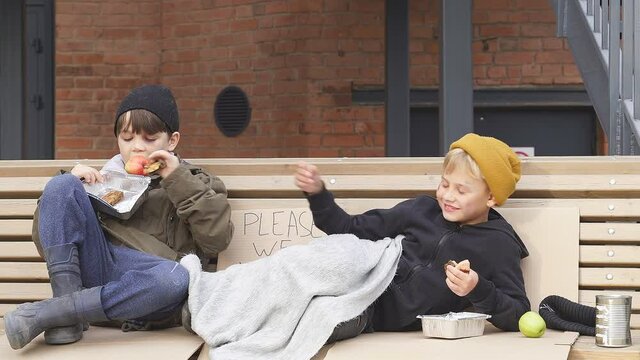 Two homeless boys sit eating food on the bench, little caucasian children, street kids are hungry, get some food by kind people.