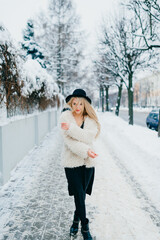 Stylish beautiful blonde girl in coat and hat posing near fence in the street