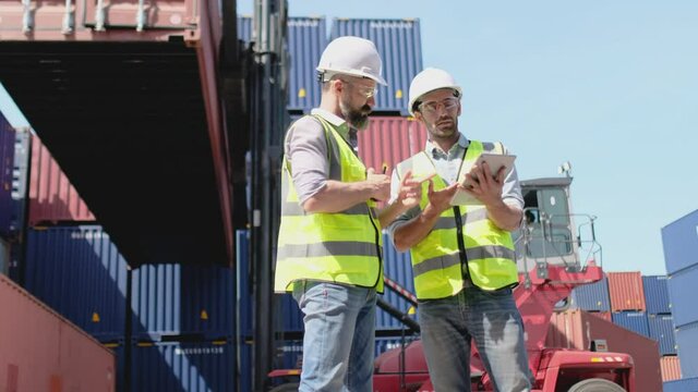 Two cargo container workers or technician men with safety uniform discuss together in front of working container truck in factory workplace area. Concept of teamwork and management system of industry.