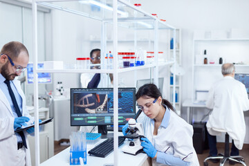 Scientist doing using microscope for medicine study. Team of researchers doing pharmacology engineering in sterile laboratory for healthcare industry with african assistant in the background.