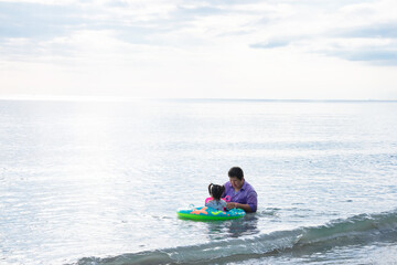 Fototapeta na wymiar Father and daughter are playing in sea. Girl sitting on green raft. Father takes care of daughter. Little child held toy in his hand. During the summer or spring. Family activities build relationships