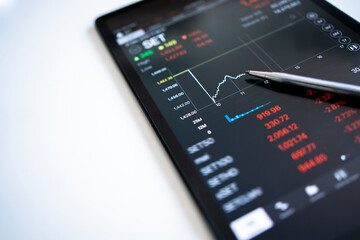 The pen points to the trader's selling price displayed as a graph falling on a tablet screen.