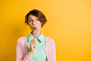 Photo of minded girl lick spoon look empty space wear specs turquoise shirt pink cardigan isolated yellow color background