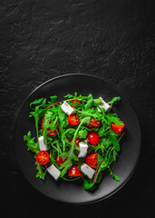 Fresh salad with tomatoes, arugula and cheese cubes on black plate on Dark grey black slate background