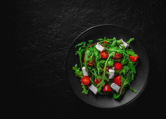 Fresh salad with tomatoes, arugula and cheese cubes on black plate on Dark grey black slate background