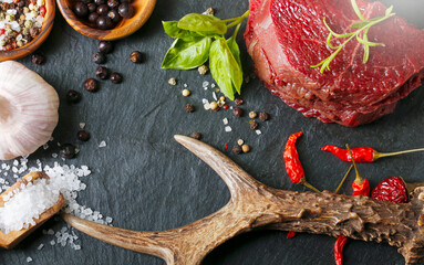 Raw steak meat from roe deer on the bridlic  chopping board  with ingredients as a sea salt, pepper ,basil,chilli and other. Roe deer antler as a decoration. - obrazy, fototapety, plakaty