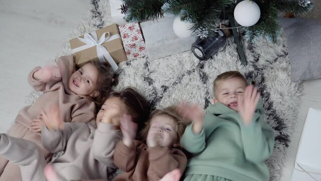 Four sweet children lay down jn a carpet near Christmas tree and waving their legs and hands. View from top