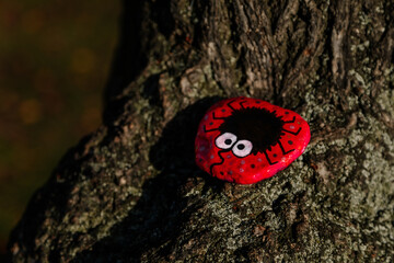 Picture of black spider with big eyes on a red stone, drawing with paints, painted stone lies on tree trunk