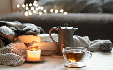 Cozy home composition with cup of tea, teapot, candlestick and books on blurred background.