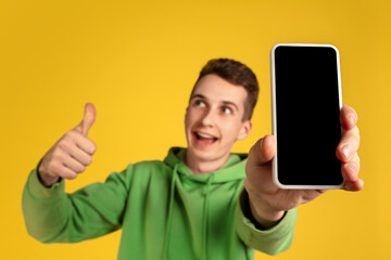 Showing blank screen. Caucasian young man's portrait on yellow studio background. Beautiful model in green outfit with cellphone. Concept of human emotions, facial expression, sales, ad, tech