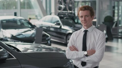 Portrait tracking of young car salesman in shirt and necktie standing with his arms crossed and looking at camera in dealership. Suitable for deepfake generation or face swap