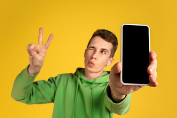 Showing blank screen. Caucasian young man's portrait on yellow studio background. Beautiful model in green outfit with cellphone. Concept of human emotions, facial expression, sales, ad, tech