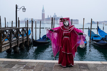 Fototapeta na wymiar Venice, Italy - February 18, 2020: An unidentified woman in a carnival costume in front of a group of gondolas and St Giorgio's Island, attends at the Carnival of Venice.