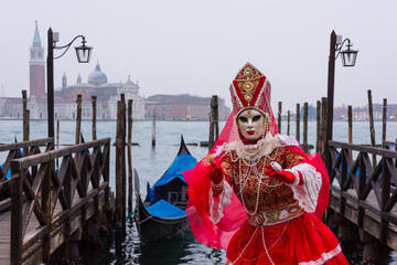 Fototapeta na wymiar Venice, Italy - February 18, 2020: An unidentified woman in a carnival costume in front of a group of gondolas and St Giorgio's Island, attends at the Carnival of Venice.
