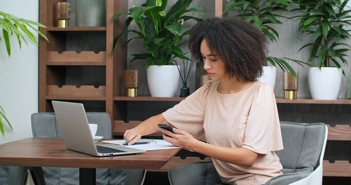 Side view of focused african american girl black curly woman female student freelancer writes on paper with pen looks into screen of mobile smart phone working remotely makes business report documents