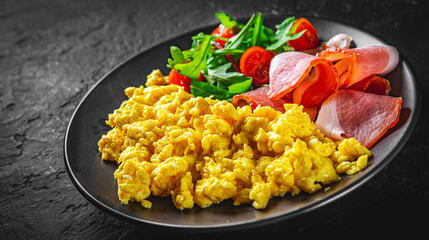scrambled eggs with prosciutto and salad on black plate on Dark grey black slate background