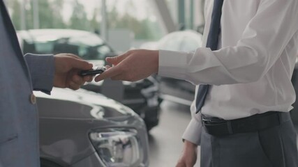 Mid-section close up of unrecognizable salesman in formal attire handing car keys to male customer and shaking hands in dealership after closing deal