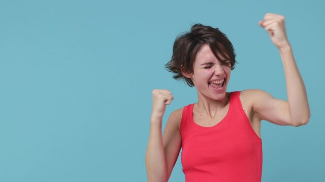 Shocked young brunette woman 20s in pink tank top posing isolated on pastel blue color background in studio. People lifestyle concept. Pointing index fingers aside doing winner gesture celebrating