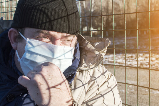 A Pandemic. A Man In A Dirty Jacket And Hat With A Medical Mask On His Face Sits At The Fence Of The Grid. Homeless People.