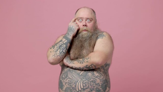 Puzzled excited fat pudge obese chubby overweight bearded man has tattooed naked bare big belly isolated on pink background studio. People lifestyle concept. Hold index finger up with great new idea