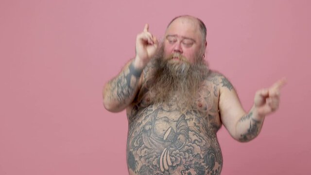 Cheerful fat pudge obese chubby overweight bearded man has tattooed naked bare big belly isolated on pink background. People lifestyle concept. Dancing clenching fists waving hands pointing fingers