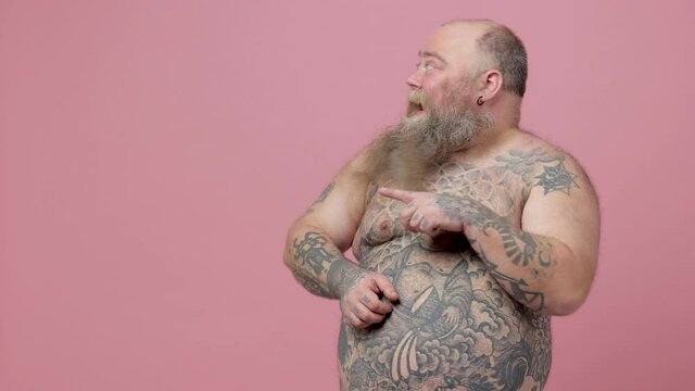 Shocked happy fat pudge obese chubby overweight bearded man has tattooed naked bare big belly isolated on pink background studio. People lifestyle concept. Pointing index finger aside clenching fists