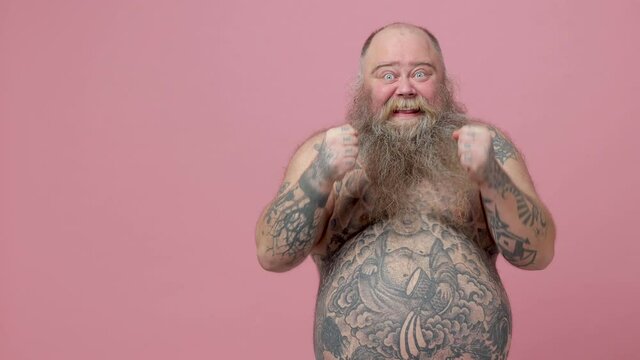 Excited fat pudge obese chubby overweight bearded man has tattooed naked big belly isolated on pink background. People lifestyle concept. Pointing camera aside doing winner gesture showing thumbs up