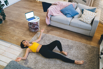 Joyful fit girl in tracksuit lying on rug and laughing during break from home workout and aerobic...