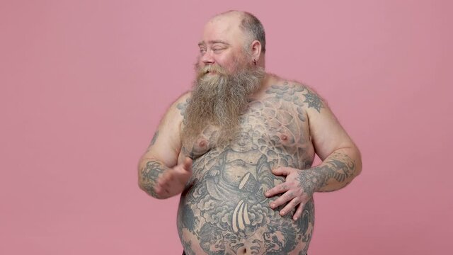 Fun fat pudge obese chubby overweight bearded man stroking slapping on tattooed naked bare big belly isolated on pastel pink background studio. People lifestyle concept. Showing thumb up like gesture