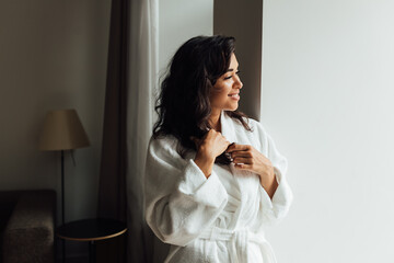 Side view of a brunette woman in a white bathrobe standing in living room and looking at a window