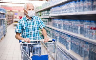 elderly european man in mask with covid protection selects bottled water in department of supermarket