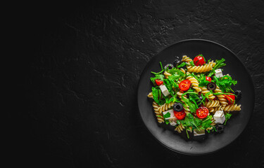 Salad with pasta, arugula, tomato, cheese and olives on black plate on Dark grey black slate background