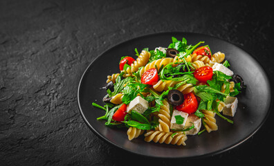 Salad with pasta, arugula, tomato, cheese and olives on black plate on Dark grey black slate background
